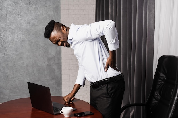 Afrykański mężczyzna trzyma się za plecy i odczuwa ból pleców i szyi po długich godzinach pracy z laptopem online w biurze Osteochondroza kręgosłupa szyjnego to zespoły korzeniowe afroamerykanina