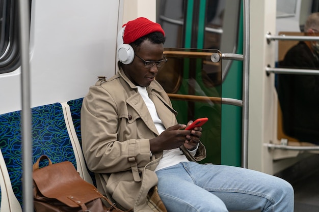 Afrykański młody człowiek w słuchawkach czyta posty słuchając muzyki za pomocą smartfona jeżdżącego w transporcie