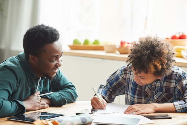 Afrykański ojciec pomaga synowi uczyć się podczas edukacji online w domu. Mężczyzna wyjaśnia materiał, a chłopiec robi notatki w zeszycie