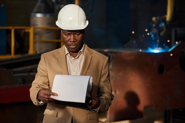 Afrykański poważny inżynier w kasku, badający plan pracy na papierach, stojąc w warsztacie w fabryce