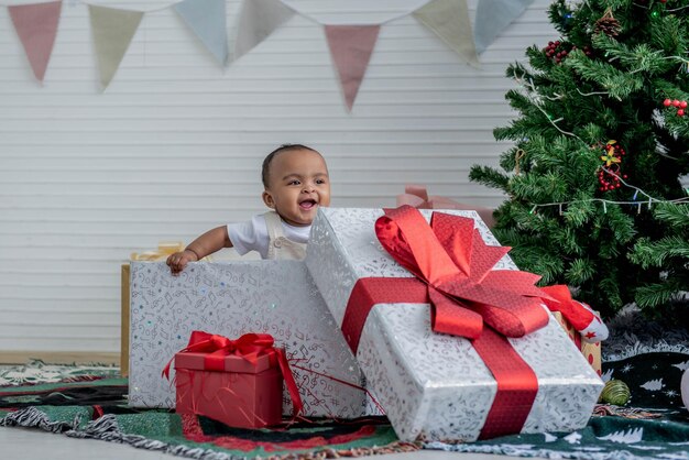 Afrykańskie dziecko, pół tajskie, pół nigeryjskie, ma 9 miesięcy, uśmiecha się i stoi w dużych pudełkach z prezentami w pobliżu drzew Bożego Narodzenia dla niemowląt i świąt Nowego Roku.
