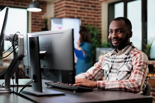 Agencja dorosły szczęśliwy uśmiechający się pracownik przy komputerze w starcie studio przestrzeni nowoczesne wnętrze. African American pracownik firmy kreatywnej siedzi przy biurku w obszarze roboczym biura, patrząc na kamery.