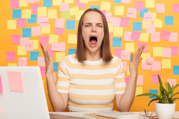 Agresywna kobieta siedząca przy stole i pracująca na laptopie przy żółtej ścianie pokrytej karteczkami samoprzylepnymi podniesione ręce trzyma szeroko otwarte usta i krzyczy, że jest zła, ma problemy w pracy