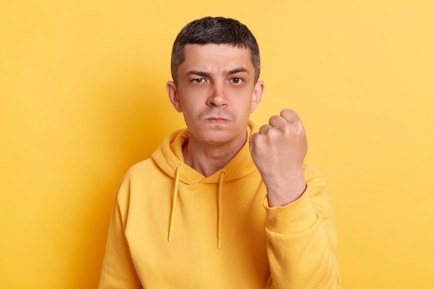 Agresywny facet Wściekła kłótnia Poważny surowy mężczyzna ubrany w swobodną bluzę z kapturem pokazujący zaciśniętą pięść kłócący się z żoną patrzącą na kamerę pozującą na białym tle na żółtym tle