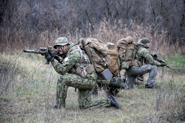 Airsoft, drużyna strajkujących, grupa komandosów poruszająca się ostrożnie po lesie, klęcząca i rozglądająca się, obejmująca towarzyszy, kontrolująca sektory. Dowódca pokazujący sygnał ręczny stop lub stop