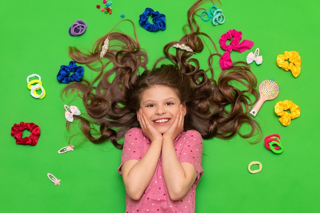 Akcesoria do włosów Szczęśliwa mała dziewczynka leży otoczona gumkami i spinkami do włosów Fryzury dla dzieci Zielone tło odizolowane