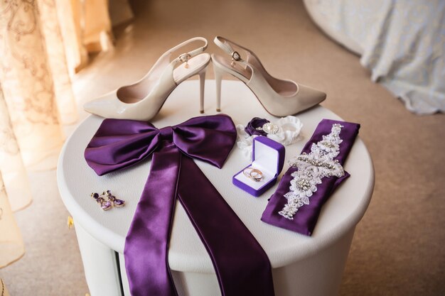 Akcesoria ślubne: buty na wysokich obcasach dla panny młodej, elementy sukni ślubnej i obrączki na pudełku z pierścionkiem na białym stole.