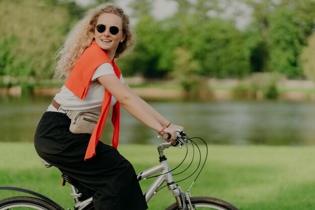 Aktywna kaukaska kędzierzawa kobieta odwraca wzrok, jeździ na rowerze sportowym, pokonuje dystanse i podróżuje na świeżym powietrzu, ubrana w codzienne ubrania pozuje w pobliżu małej rzeki z zielonymi drzewami prowadzi zdrowy tryb życia