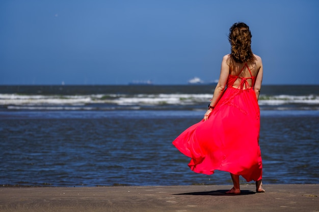 Aktywna kobieta w czerwonej sukience chodzi po plaży z włosami płynącymi na horyzoncie nad wodą