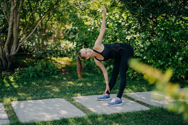 Zdjęcie aktywna modelka fitness ćwiczy na świeżym powietrzu pochyla się do stóp unosi ramię przygotowuje mięśnie przed wyzwaniami treningu cardio sama ubrana w strój sportowy pozuje w koński ogon w zielonym parku w słoneczny dzień