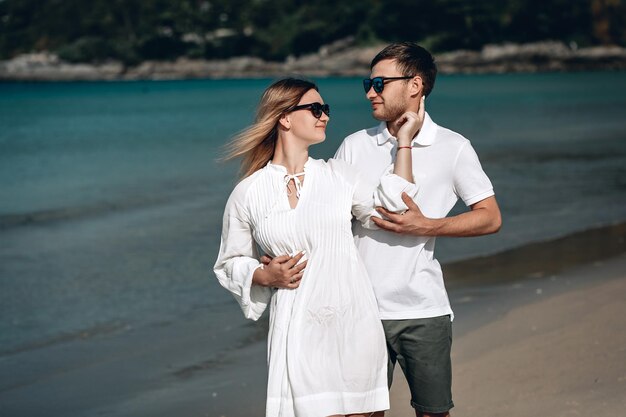 Aktywna modna młoda para w białych strojach stoi na plaży i trzyma się za ręce. Piękna blondynka z czerwoną nitką na ramieniu i mężczyzna są na wakacjach. Phuket. Tajlandia