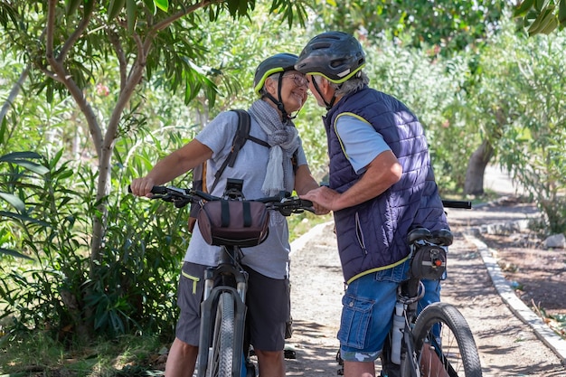 Aktywna Para Seniorów Rasy Kaukaskiej Z Elektrycznymi Rowerami Stojącymi Na Zewnątrz W Parku Wita Się Całując Dwóch Uśmiechniętych Seniorów W Kasku Rowerowym Na łonie Natury, Cieszących Się Zdrowym Stylem życia