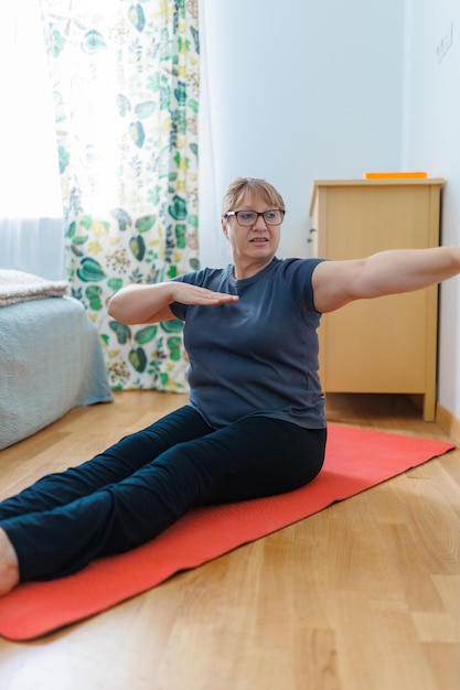 Aktywna starsza kobieta robi ćwiczenia abs na macie do jogi w domu i uśmiecha się do kamery