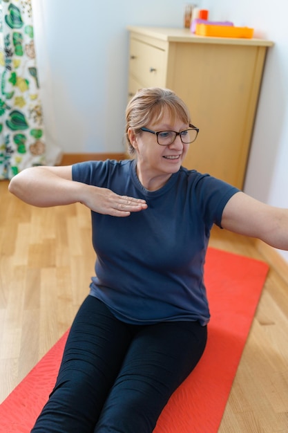 Aktywna starsza kobieta robi ćwiczenia abs na macie do jogi w domu i uśmiecha się do kamery