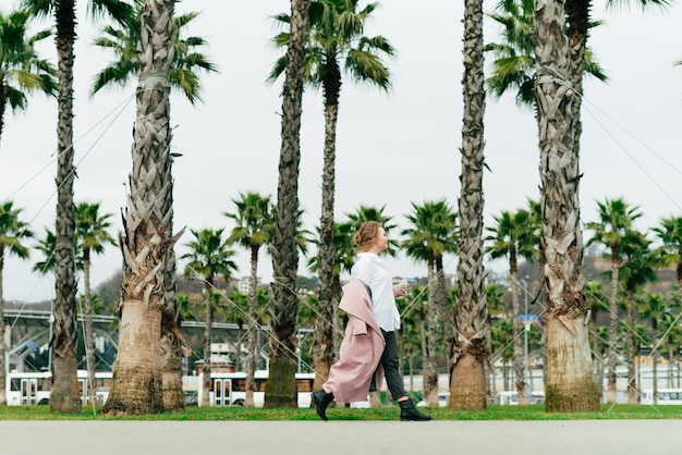 Aktywna stylowa dziewczyna w białej koszuli spaceruje obok wysokich zielonych palm, ciesząc się wiosną
