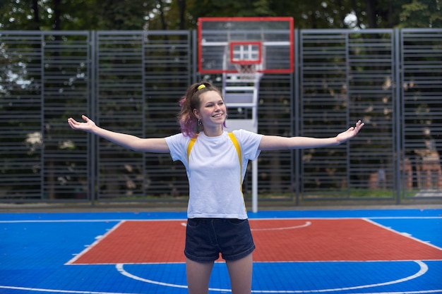 Zdjęcie aktywna uśmiechnięta nastolatka, modna z fioletowymi włosami, na boisku do koszykówki ulicznej z podniesionymi rękami. emocja szczęścia, wakacji, zabawy