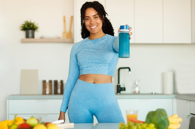 Aktywne sporty w domu i pragnienie Szczęśliwa czarna kobieta w odzieży sportowej trzymająca butelkę z wodną pozycją w kuchni