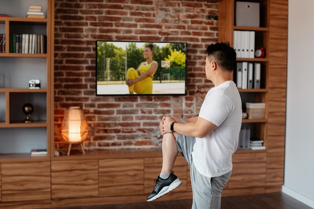 Aktywny azjatycki dojrzały mężczyzna ćwiczący w domu i oglądający program ćwiczeń fitness w telewizji, wykonujący ćwiczenia rozgrzewające