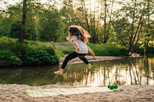 Zdjęcie aktywny mały sportowiec z długimi włosami skaczący wysoko na sportowym dywanie na piaszczystej plaży w pobliżu jeziora dziewczyna trenująca w przyrodzie