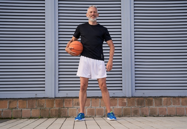 Aktywny Mężczyzna W średnim Wieku W Odzieży Sportowej, Trzymający Piłkę Do Koszykówki I Uśmiechający Się Do Kamery Podczas Stania