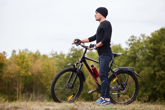 Aktywny sportowiec rowerzysta ubiera czarny dres stojący z rowerami na wzgórzu w pobliżu zielonych drzew, ciesząc się widokiem przed nim