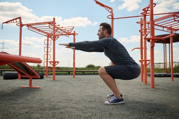 Aktywny Sportowiec W Stroju Lekkoatletycznym Wykonujący Przysiady Podczas Treningu Na Boisku Zewnętrznym. Młody Człowiek Uprawiania Sportu Na Letnim Boisku. Koncepcja Zdrowego I Aktywnego Stylu życia