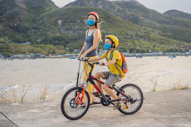 Aktywny szkolny chłopiec i jego mama w masce medycznej i kasku ochronnym jeżdżący na rowerze z plecakiem