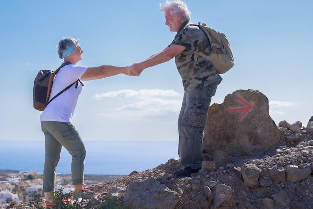 Aktywny uśmiechający się starszy para w plenerowej wycieczce piesze wycieczki w góry z plecakiem, ciesząc się zdrowym stylem życia Brodaty mężczyzna pomaga kobiecie wspinać się po horyzont nad wodą na tle