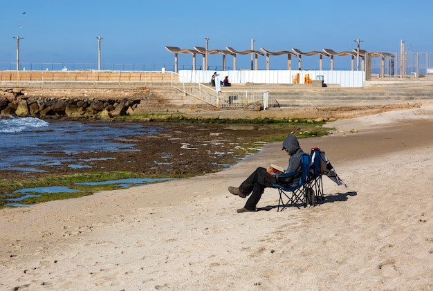 Amerykanin relaks na czytaniu książki na plaży podczas zachodu słońca. Rosyjski człowiek czytanie książki w leżaku z widokiem na morze. przystojny brodaty mężczyzna rasy kaukaskiej w okularach przeciwsłonecznych, czapce i kurtce z kapturem