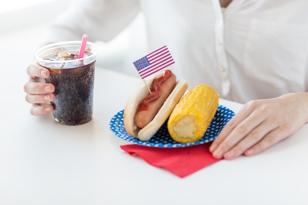 amerykański dzień niepodległości, celebracja, patriotyzm i koncepcja świąt - zbliżenie rąk kobiety z hot dogiem i kukurydzą trzymającą napój coca cola w plastikowym kubku na imprezie 4 lipca