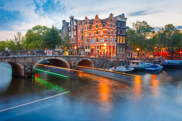 Amsterdamski kanał, most i typowe domy, łodzie i świetlisty ślad z łodzi podczas porannej godziny zmierzchu, Holandia, Holandia.