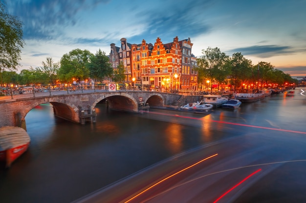 Amsterdamski kanał, most i typowe domy, łodzie i świetlisty ślad z łodzi podczas porannej godziny zmierzchu, Holandia, Holandia.