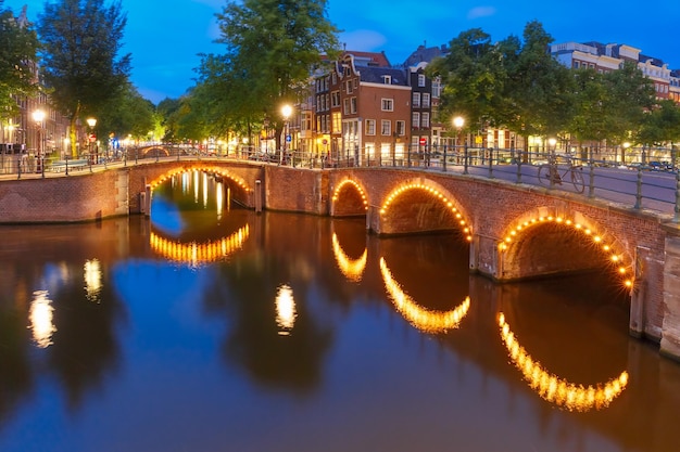 Amsterdamski most nad kanałem i typowe domy łodzie i rowery podczas wieczornego zmierzchu blue hour holla