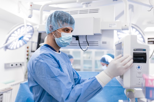 Zdjęcie anestezjolog pracujący na sali operacyjnej noszący sprzęt ochronny sprawdzający monitory podczas uspokajania pacjenta przed zabiegiem chirurgicznym w szpitalu