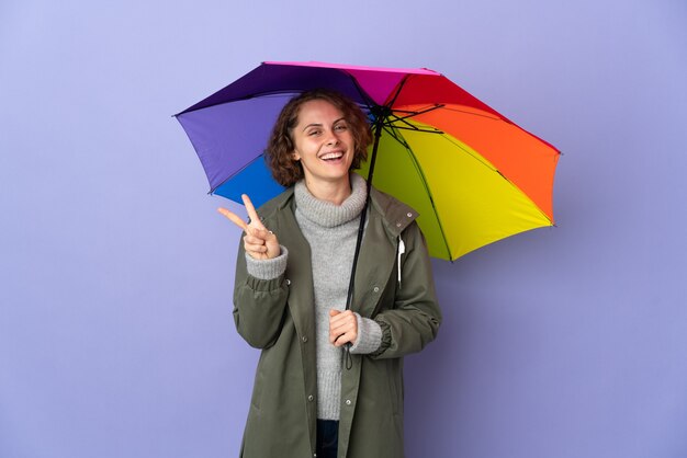 Angielska kobieta trzyma parasol na białym tle na fioletowej przestrzeni, uśmiechając się i pokazując znak zwycięstwa