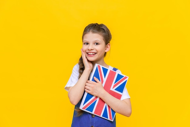 Angielski dla dzieci Mała dziewczynka trzyma w rękach podręcznik z angielską flagą Edukacja językowa dla dzieci Żółte tło izolowane