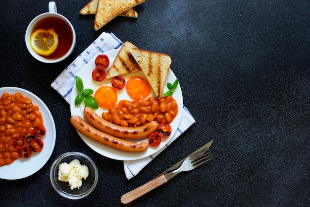 Angielskie śniadanie ze smażonymi kiełbasami, fasolą, pieczarkami, jajkami sadzonymi, grillowanymi pomidorami cherry. Podawany z filiżanką herbaty z cytryną, tostem chlebowym i masłem. Czarne tło, widok z góry, lato