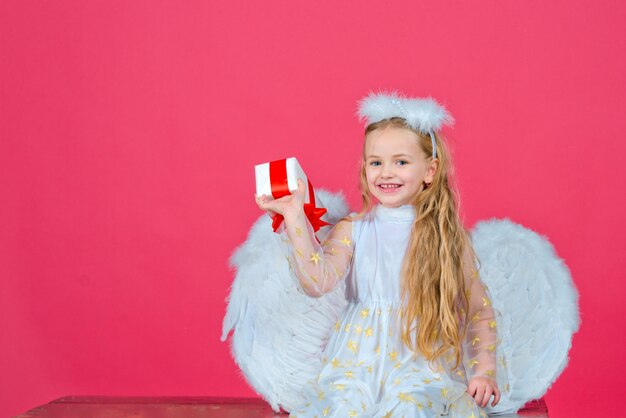 Anielskie dzieci, szczęśliwa wróżka z prezentem, portret studyjny. Mały aniołek blondynka z białymi skrzydłami trzyma prezent. Cute dziewczynka dziecko anioł ze skrzydłami anioła, na czerwonym tle. Walentynki.