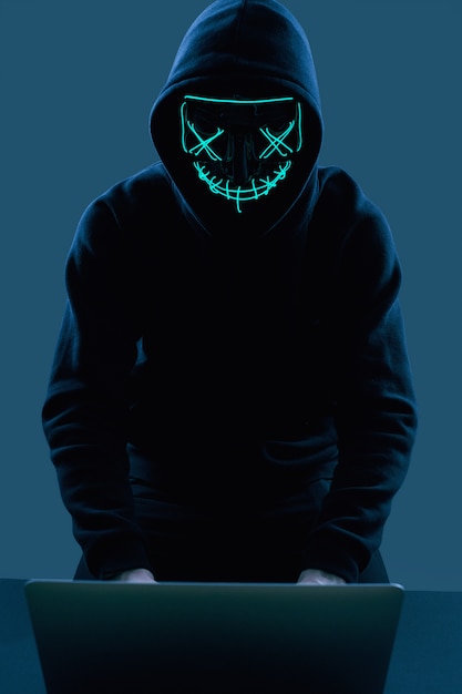 Anonimowy mężczyzna w czarnej bluzie i neonowej masce włamujący się do komputera