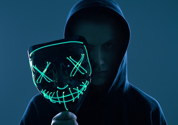 Anonimowy mężczyzna w czarnej bluzie z kapturem chowający twarz za neonową maską