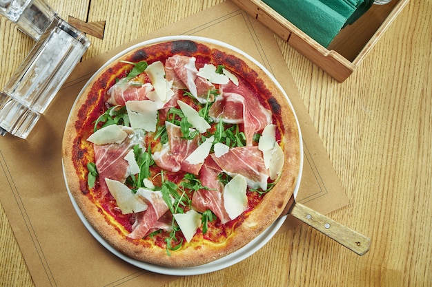 Apetyczna pieczona pizza z szynką parmeńską, parmezanem i rukolą z chrupiącą skórką na drewnianym stole. Ustawienie stołu w restauracji. Widok z góry