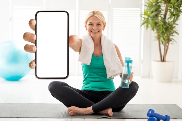 Zdjęcie aplikacja fitness starsza kobieta siedzi na macie do jogi i pokazuje pusty smartfon