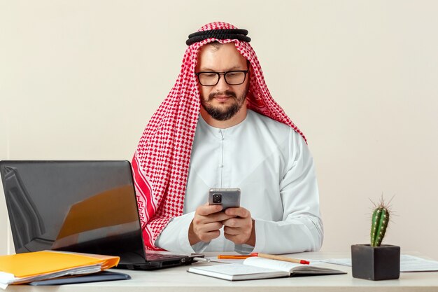 Arab, biznesmen, szejk w narodowym stroju arabskim pracuje przy stole w biurze. Inwestycje, biznes, praca przez internet, umowy online.