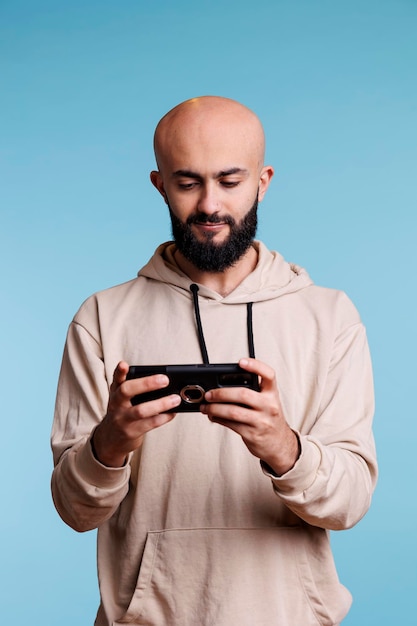 Arab korzystający z aplikacji na smartfony i grający w gry wideo podczas spędzania wolnego czasu. Młoda uśmiechnięta osoba trzymająca telefon komórkowy w trybie pionowym i oglądająca wideo online