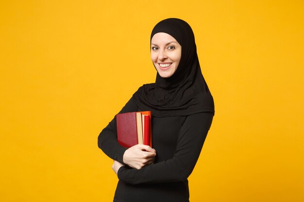 Arabian muzułmańskich student dziewczyna w hidżab czarne ubrania posiada książki na białym tle na żółtą ścianę portret. Ludzie religijny styl życia, edukacja w koncepcji liceum. .