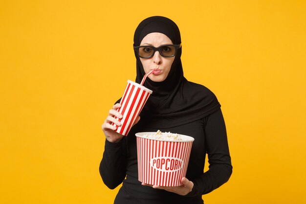 Arabska kobieta muzułmańska w hidżab czarne ubrania 3d okulary imax oglądać film film trzymać popcorn, filiżankę sody na białym tle na żółtą ścianę portret. Koncepcja życia ludzi. .