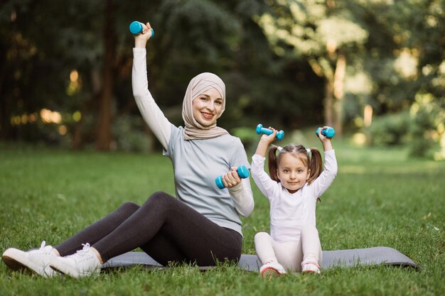 Arabska matka w hidżabie i 4-letnia córka siedzi na macie w parku i podnosi hantle
