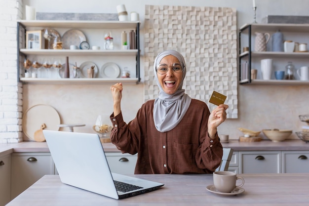 Arabska młoda kobieta siedzi w domu w kuchni za pomocą laptopa i karty kredytowej, zadowolona z ręki