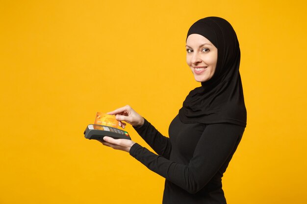 Arabska Muzułmanka W Czarnych Ubraniach Hidżab Trzyma Terminal Płatniczy W Celu Przetwarzania I Uzyskiwania Płatności Kartą Kredytową Na żółtej ścianie. Koncepcja życia Religijnego Ludzi.