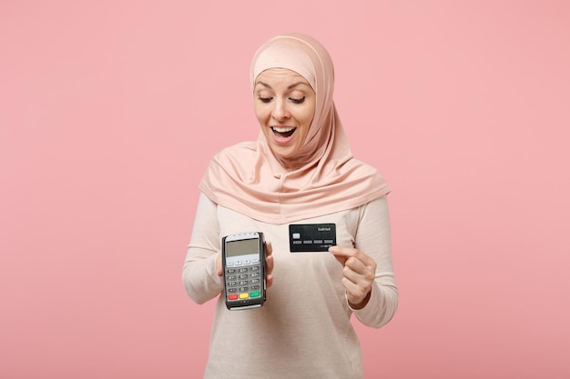 Arabska Muzułmanka w jasnych ubraniach hidżab pozowanie na białym tle na różowym tle. Koncepcja życia religijnego ludzi. Makieta miejsca na kopię. Przytrzymaj terminal płatniczy, aby przetworzyć, uzyskać płatności kartą kredytową.